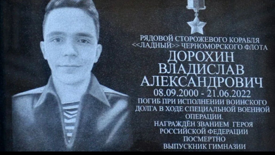 A granite memorial plaque on a school wall with fresh red carnations flowers below it, daylight, respect